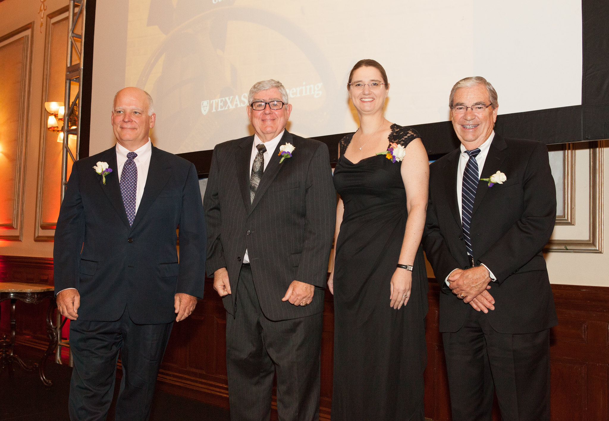 Group of four alumni honorees standing onstage smiling, backlit by large projector screen.