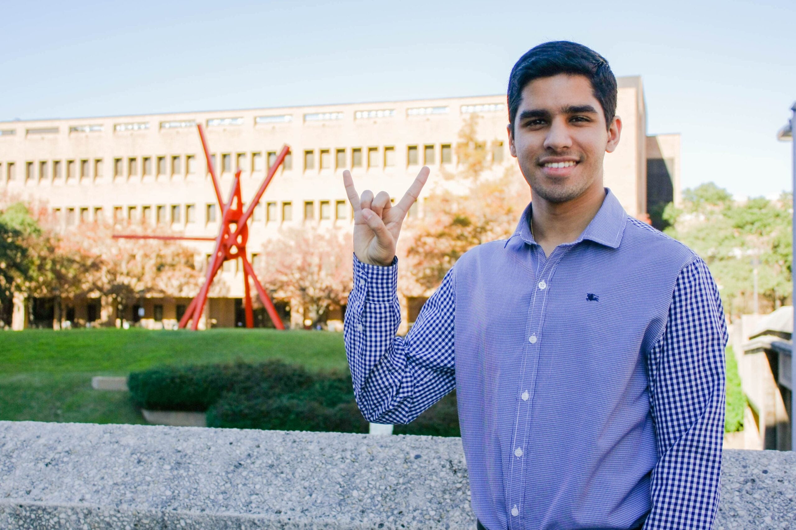 Karan Jerath smiling and doing hook 'em horns hand sign in front of Clock Knot sculpture on UT Austin campus.