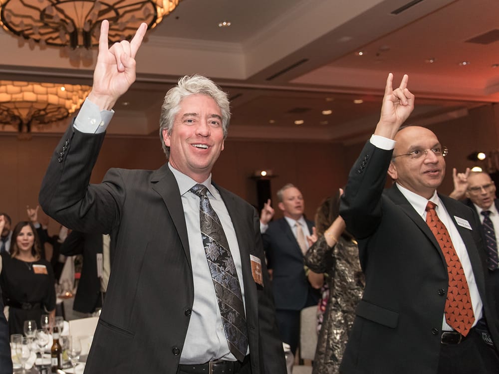 alumni at an event wearing suits and making hook 'em gesture with their hands