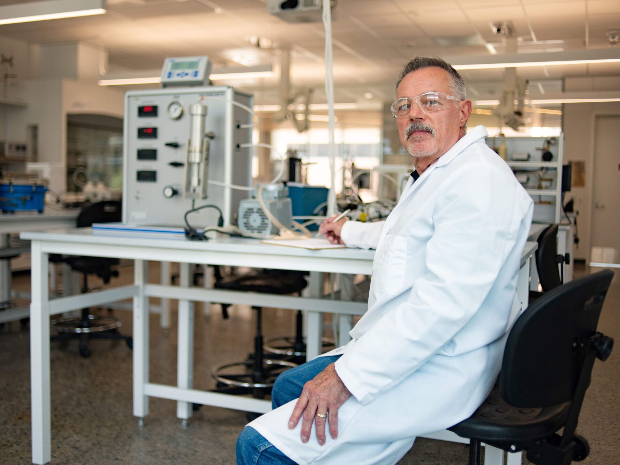 Glen Baum wearing lab coat and safety goggles, sitting at table in lab.