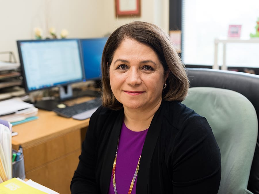 Mojdeh Delshad smiling while sitting at a desk in front of books and a computer monitor.