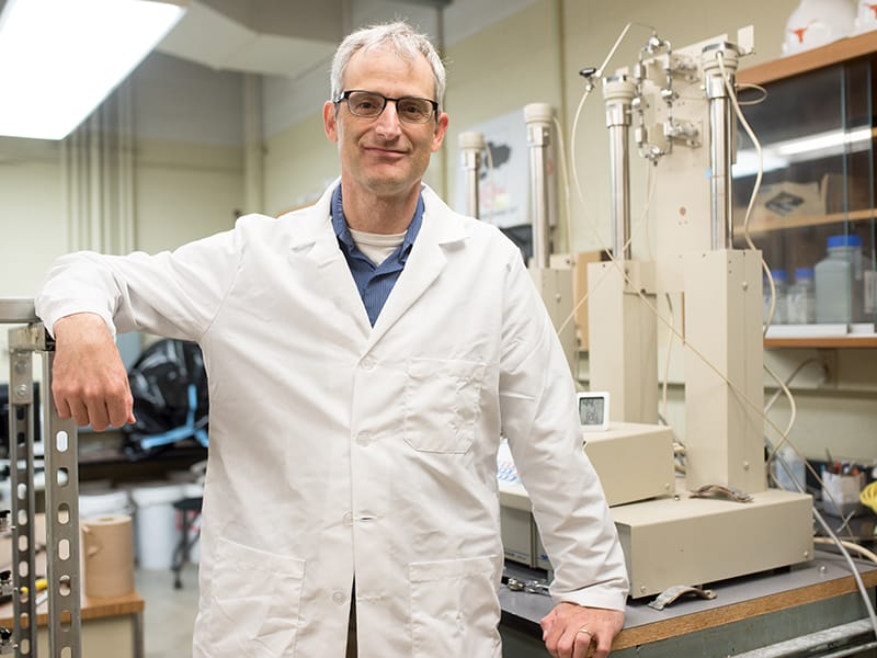 David DiCarlo resting his arm on large machine while wearing lab coat in research lab.
