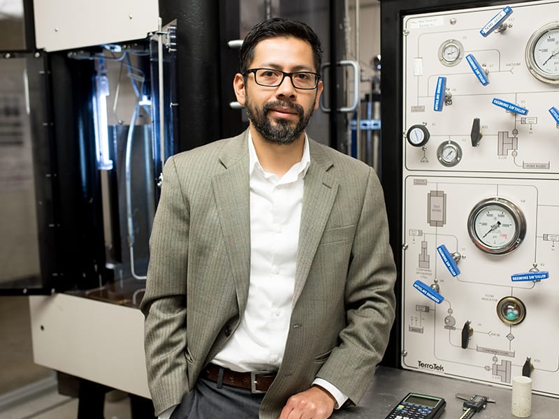 Nicolas Espinoza wearing formal suit jacket, leaning against railing in front of large laboratory equipment and smiling.