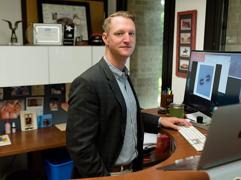 John Foster standing behind his desk with computer screens projecting research graphics.