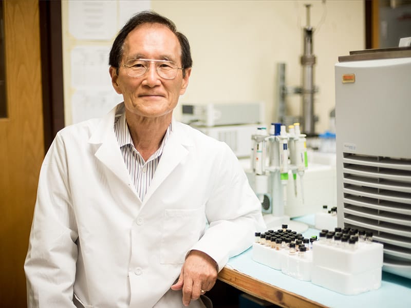 Chun Huh leaning against desk with microscope and research equipment on it while wearing lab coat.