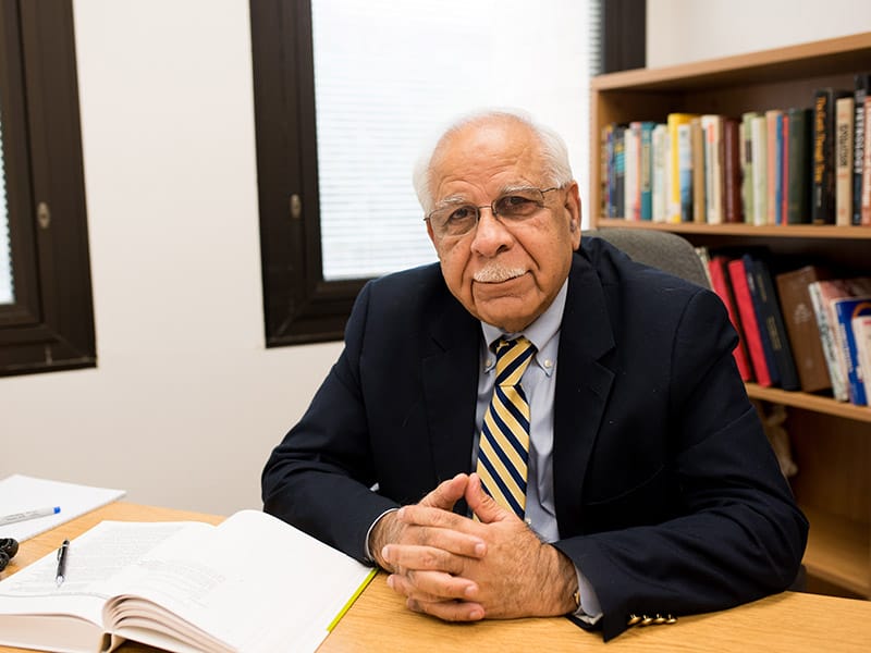 Krishan Malik sitting at desk with hands folded in front of open book.