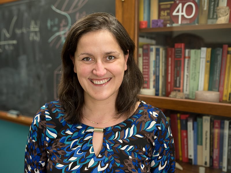 Masa Prodanovic smiling in front of bookcase and chalkboard.