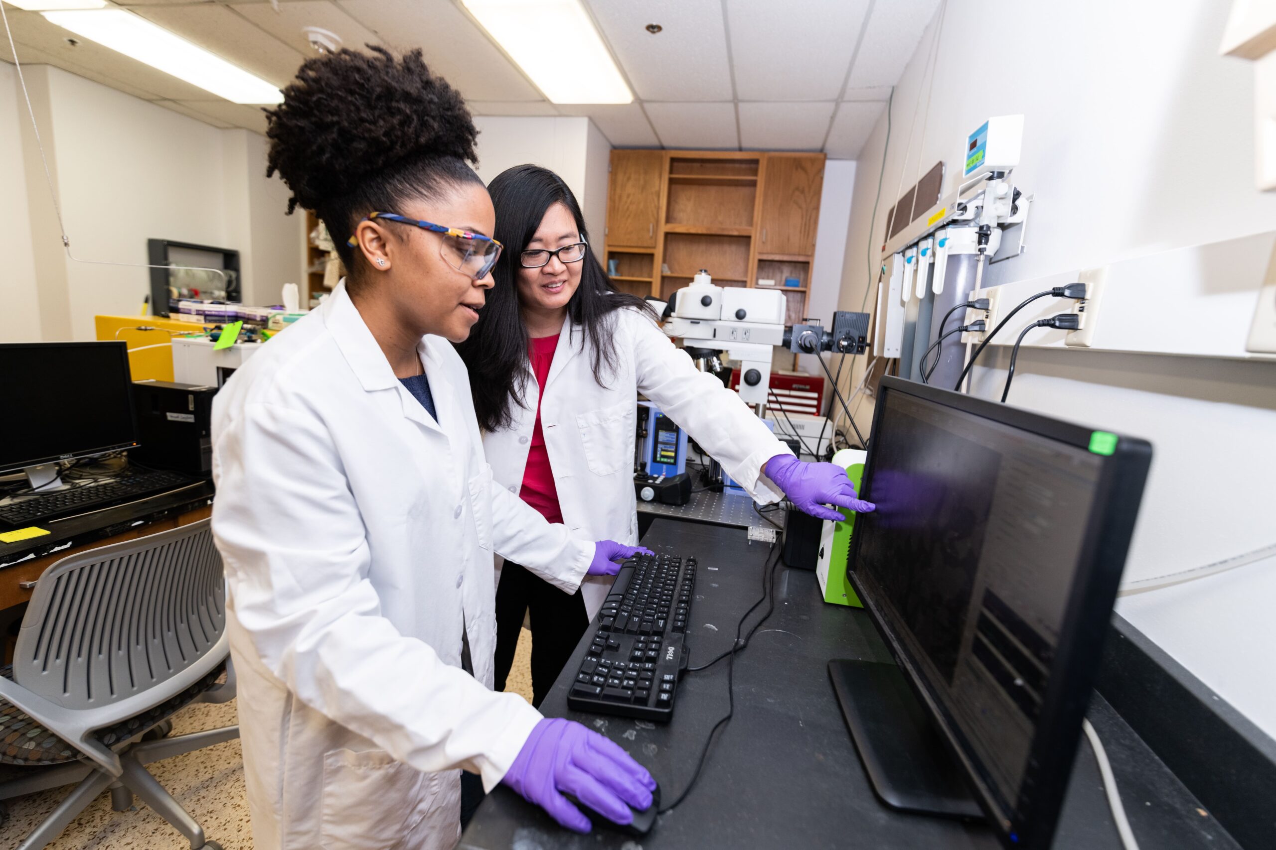 Professor Wen Song and PGE graduate student wearing lab coats and looking at computer screen; Song points at monitor.