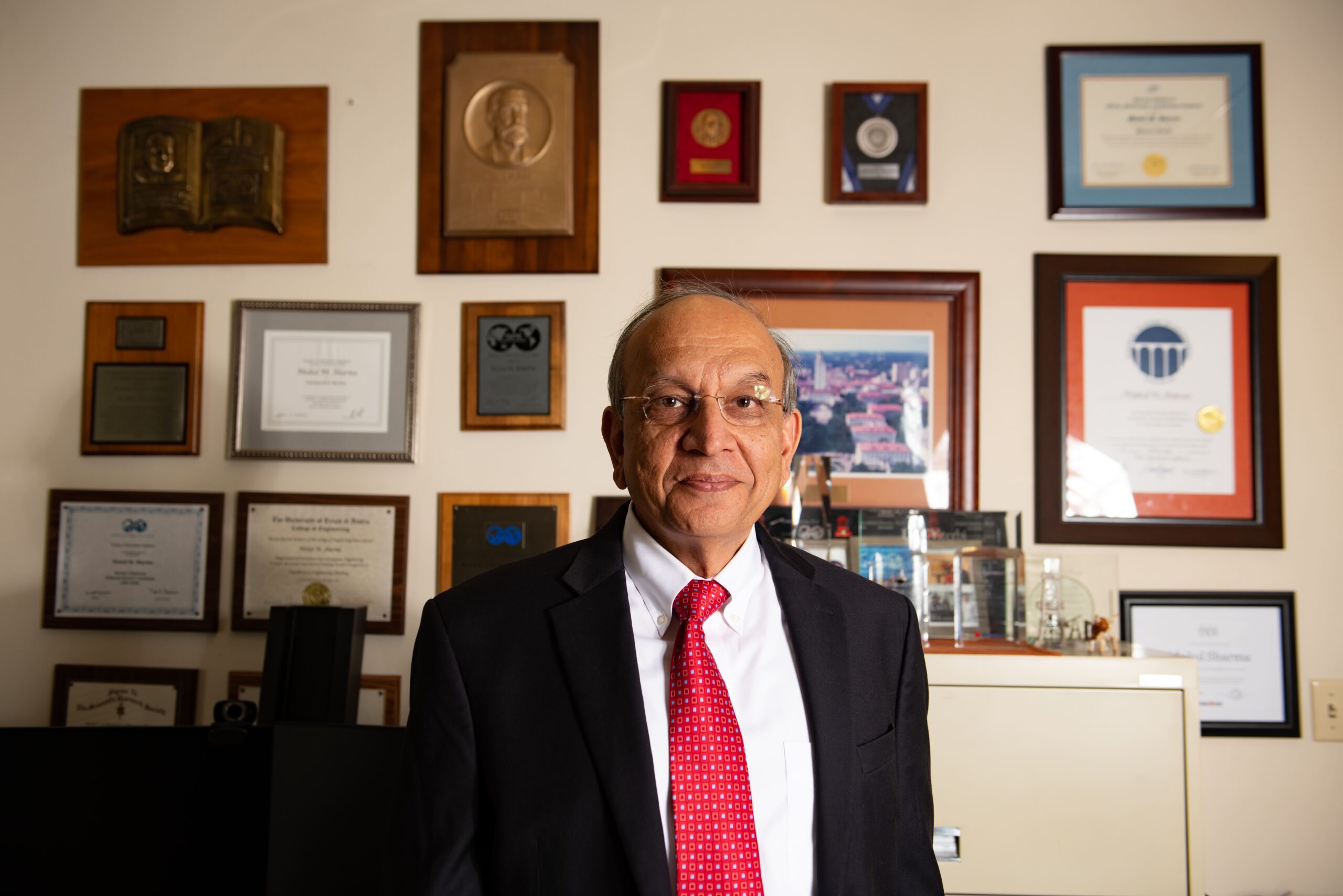 Texas Engineer Mukul Sharma smiling in office in front of wall of framed degrees and awards.