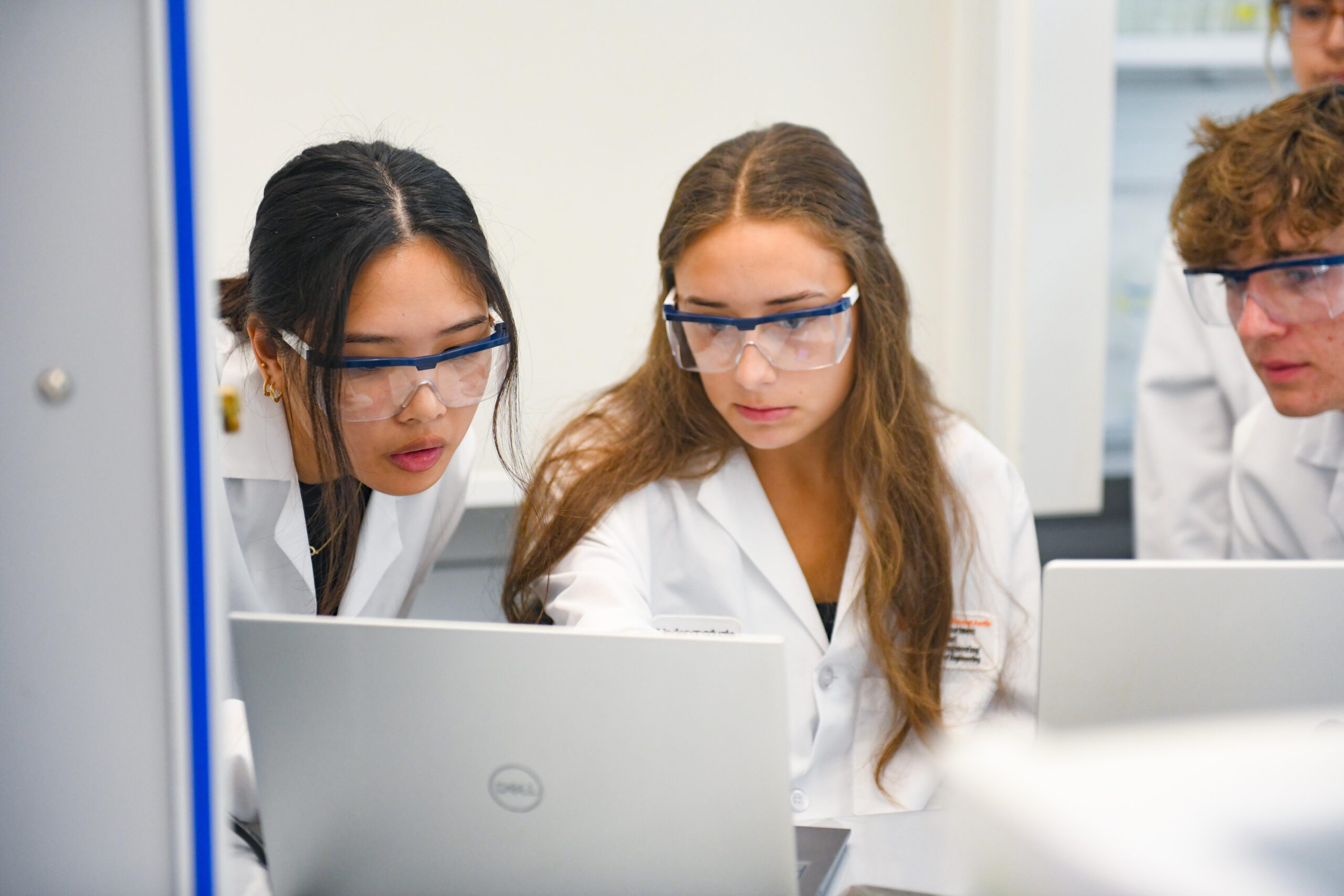 Researchers looking at laptop in lab while wearing lab coats and safety goggles.