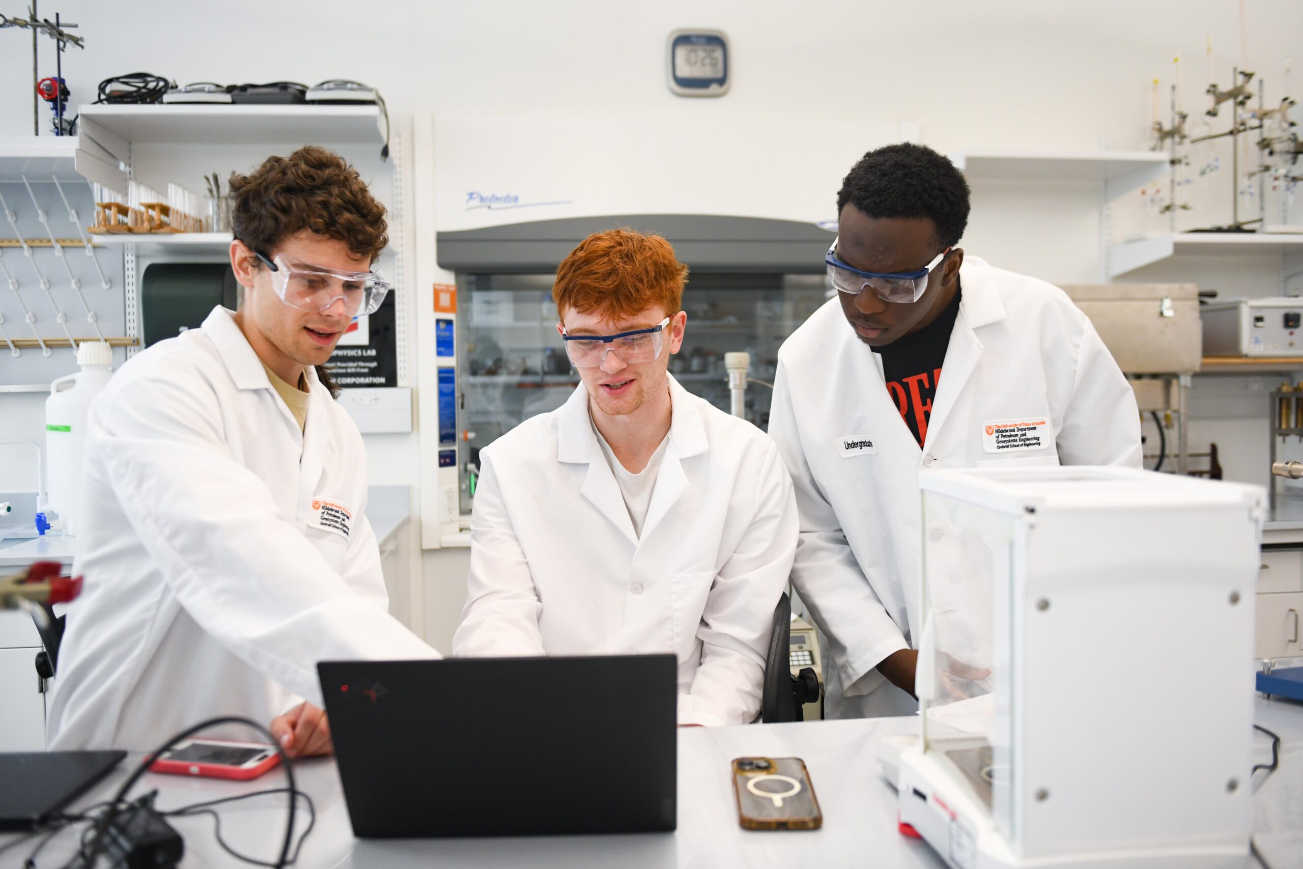 Student researchers wearing lab coats and safety goggles gathered around laptop in research lab.