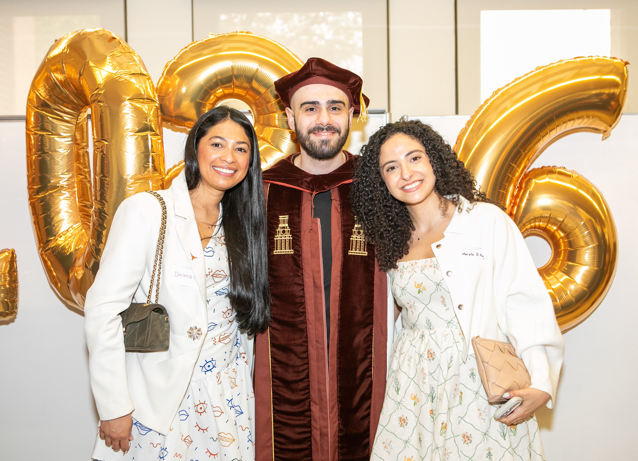 UT PGE student wearing graduation robes and regalia smiling with family in front of 2026 balloon arrangement at Graduation Reception event.
