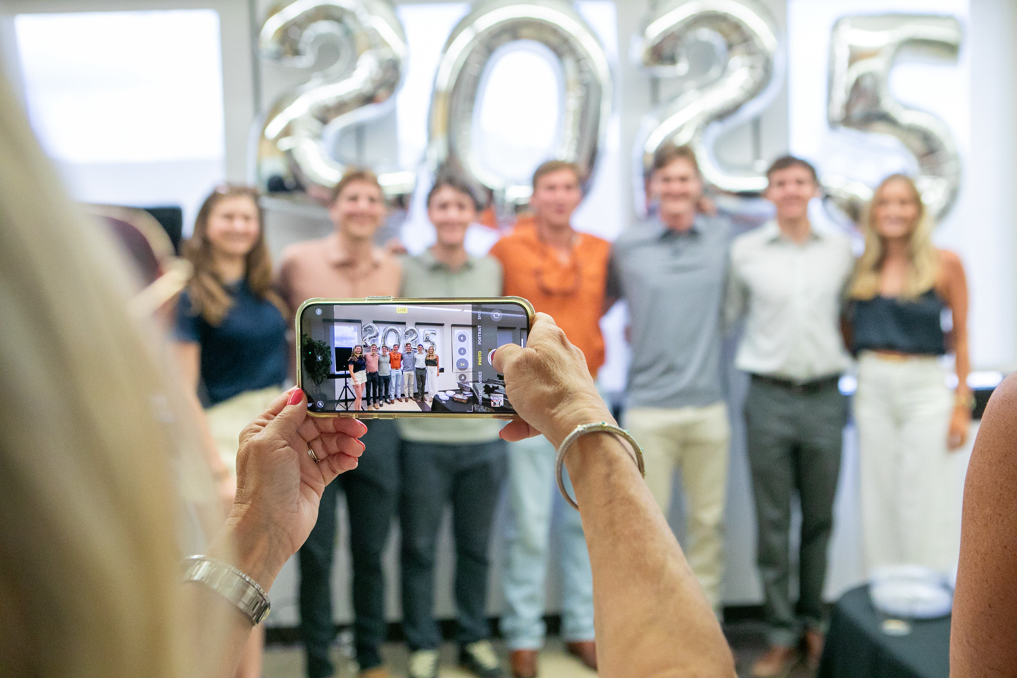Woman taking photo with iPhone of group of UT PGE students in front of 2025 ballon arrangement.