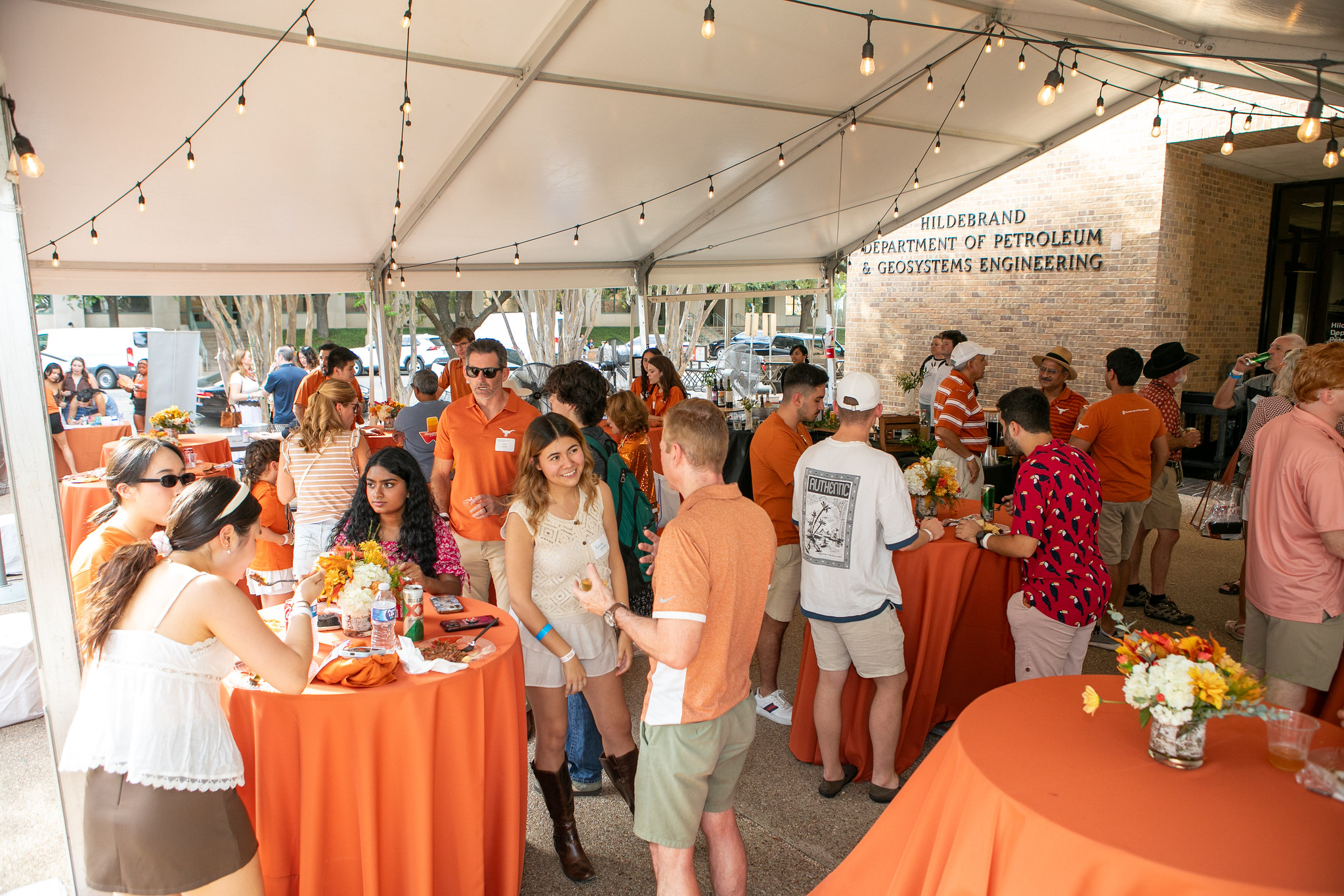 UT PGE alumni gathered under tent and talking at tables with refreshments in front of PGE building on UT Austin campus.