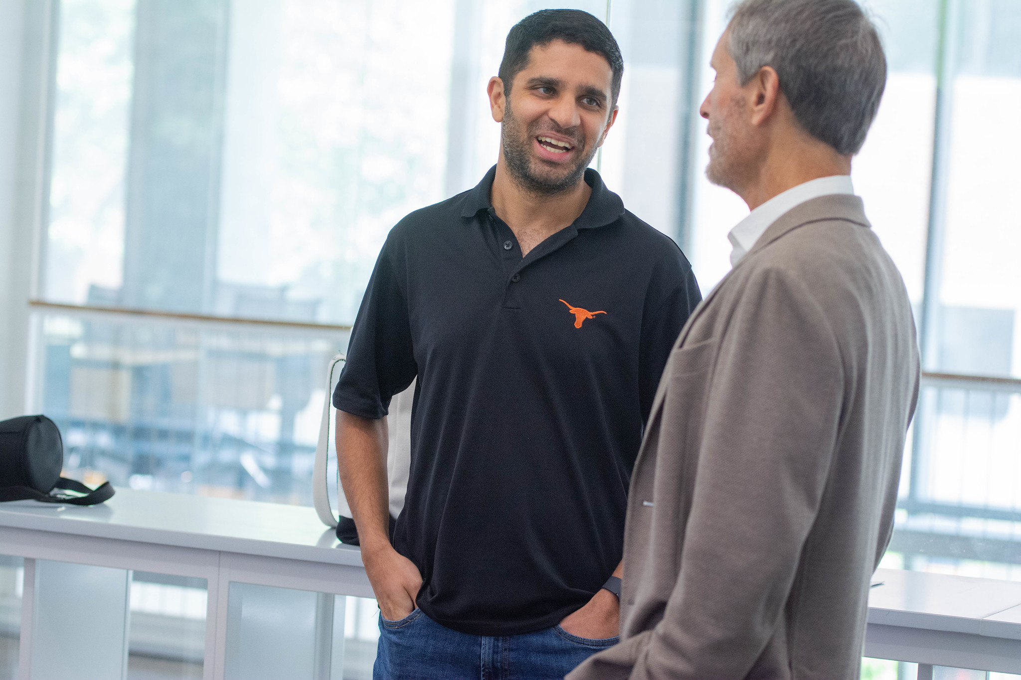 Two men talking in well-lit room with windows. One wears a suit and tie and the other wears a UT polo shirt with the UT Austin logo.