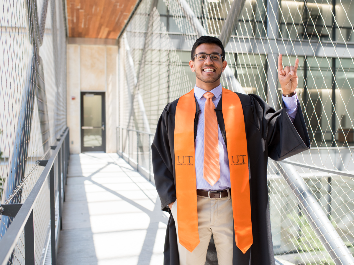 PGE student standing on a bridge, smiling and doing hook 'em horns hand sign while wearing graduation regalia.