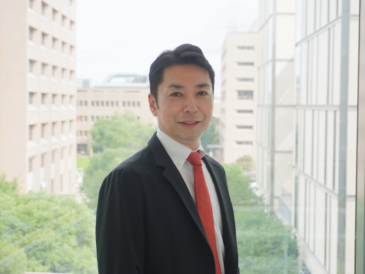 Ryosuke Okuno smiling in front of large window overlooking trees and buildings on UT Austin campus.