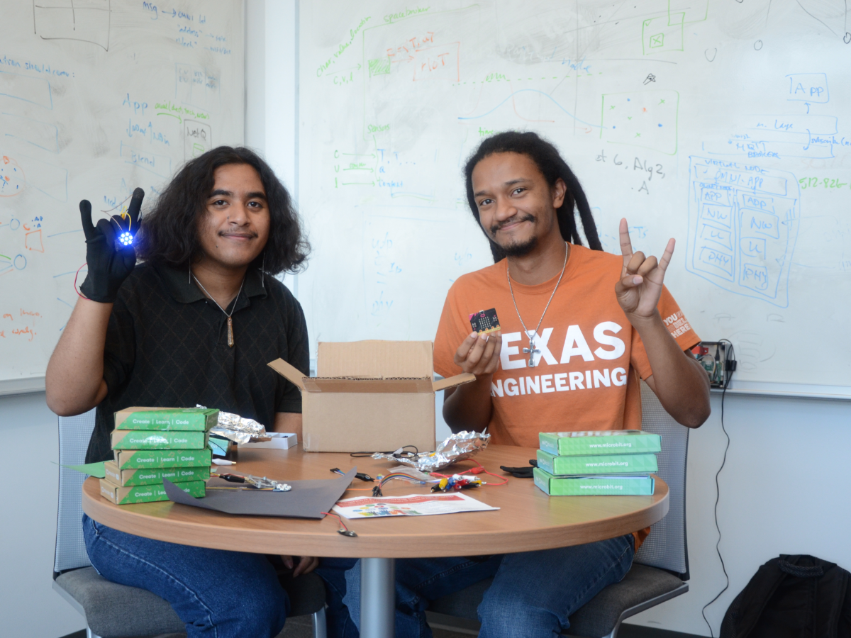Two students hold their hands up in a hook em while work on an electrical device