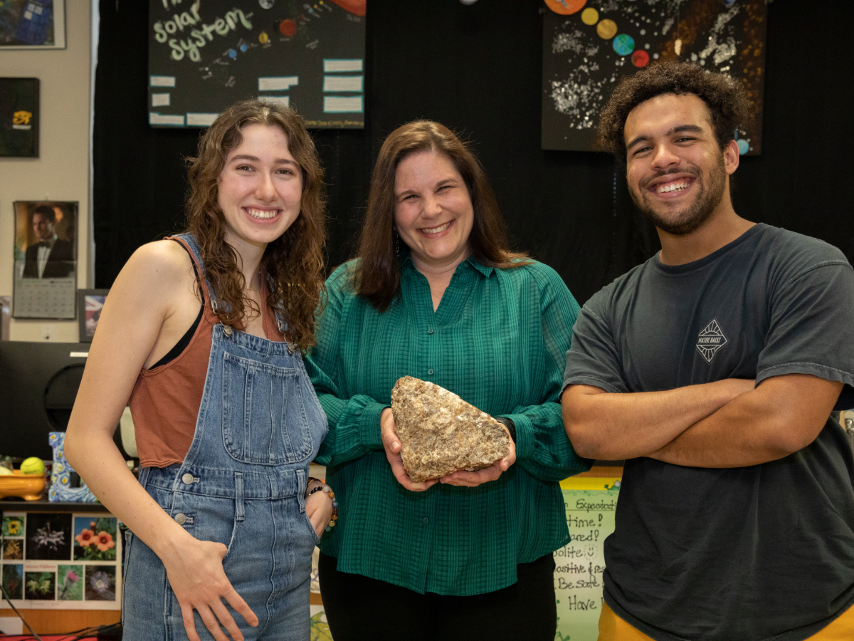 Sabrina Ewald holding large rock and smiling next to two students.
